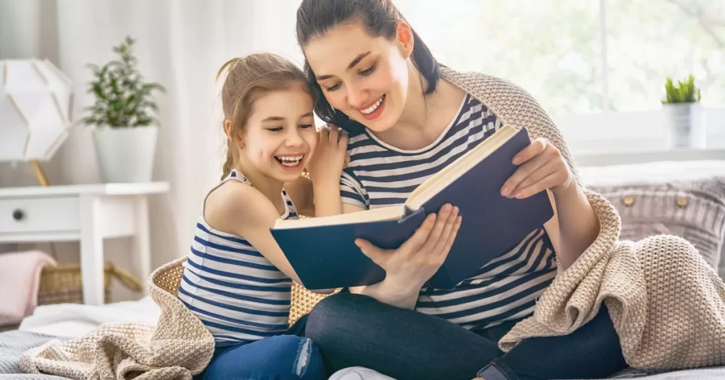 A mother and daughter sitting together, smiling and enjoying reading a book in a cozy, brightly lit room, creating a warm and inviting scene.