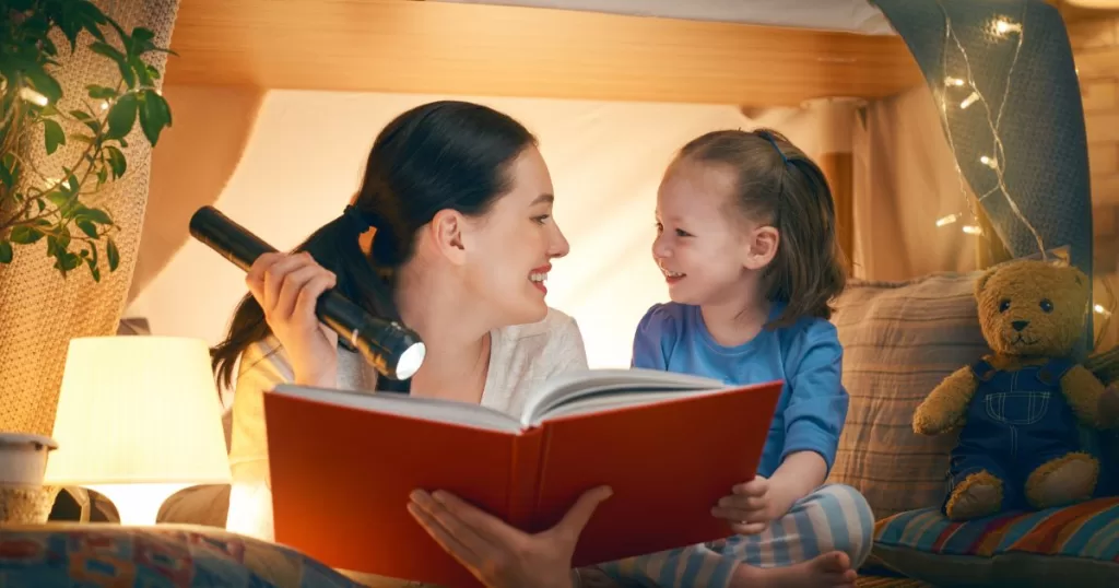 A mother and her young daughter reading a book with a flashlight in a cozy indoor tent setup, surrounded by soft lights, creating a magical and intimate reading atmosphere.