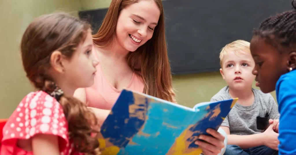 A teacher is reading a colorful book to a group of young children in a classroom. The children are gathered around, listening attentively, with expressions of curiosity and engagement. The scene reflects an interactive and supportive learning environment.