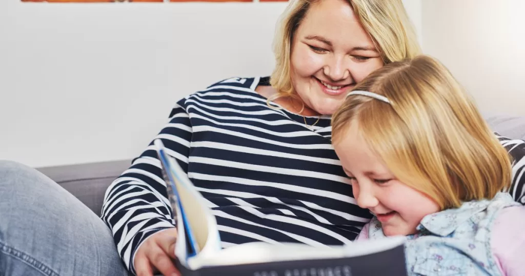 A mother and her young daughter are sitting together on a couch, smiling and enjoying a book. The mother looks on warmly while the daughter laughs, creating a joyful and cozy reading moment that highlights their bond and shared enjoyment of reading.